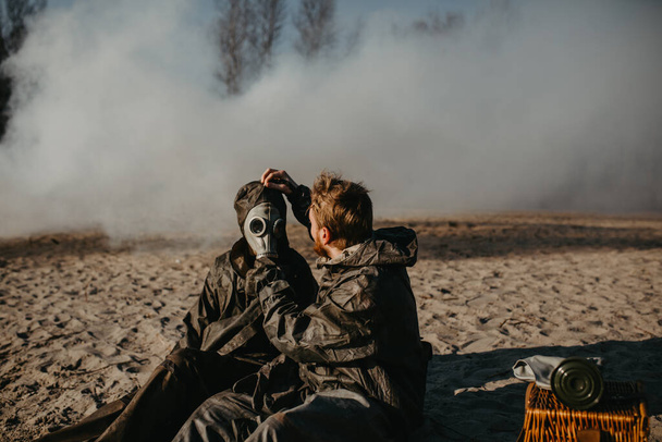 Couple in love sits at outing in NBC protective suits and removes gas masks on smoke background. Concept of a preventive measures and protection for apocalypse, global pollutions of environment and pandemic. - Photo, Image