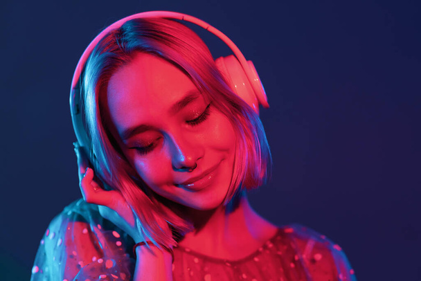 Mysterious hipster teenager listening to music with headphones. Portrait of millennial pretty girl with short hairstyle with neon light. Dyed blue and pink hair. - Fotografie, Obrázek