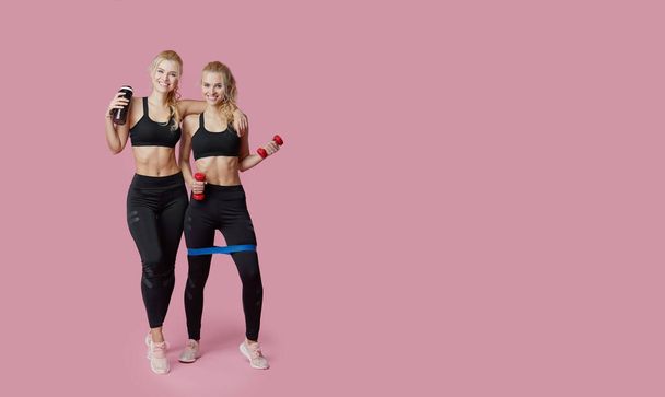 Athletic women twins practicing squat exercise with fitness expander in the studio. Girls in sportswear exercising on a pink background. Space for text - Foto, immagini