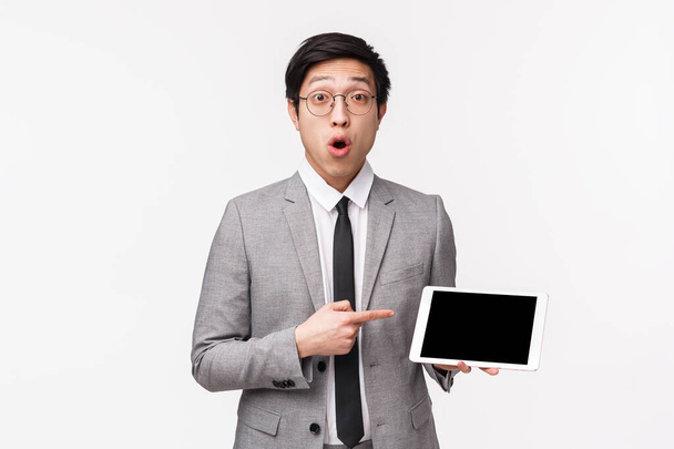 Retrato en la cintura de un joven asiático impresionado y curioso con traje gris y gafas, haciendo preguntas sobre algo que vio en Internet, apuntando a la pantalla digital de la tableta, mirar la cámara intrigada
 - Foto, imagen