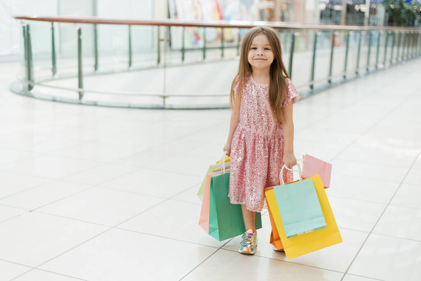 モール内の小さな幸せな女の子の肖像画。手に多色のバッグを持ったピンクのドレスを着た笑顔の女の子が店の窓を見て歩いている. - 写真・画像