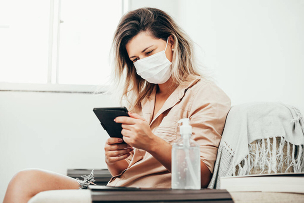 Coronavirus. Covid-19. Woman in quarantine wearing protective mask reading e-book in the living room. Bottle of alcohol gel in the foreground. - Foto, Imagem