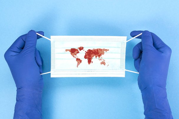 Wegwerp medische masker met de contour van de continenten van de planeet in de handen van een arts op een blauwe achtergrond, pandemische concept. KOVID-19, coronavirus. De wereldwijde epidemie van de ziekte. - Foto, afbeelding
