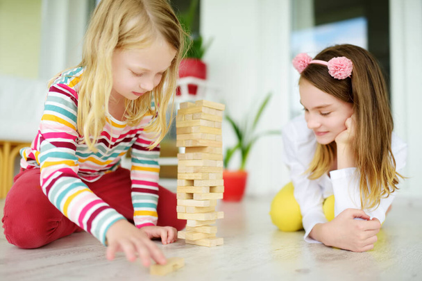 Two cute sisters playing wood blocks tower game at home. Kids practicing their physical and mental skills. Fun board games for family leisure. Stay at home activity for kids. - Foto, immagini