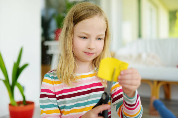 Young girl cutting colorful paper with scissors at home. Creative kid doing crafts. Education and distance learning for kids. Homeschooling during quarantine. Stay at home entertainment. - 写真・画像