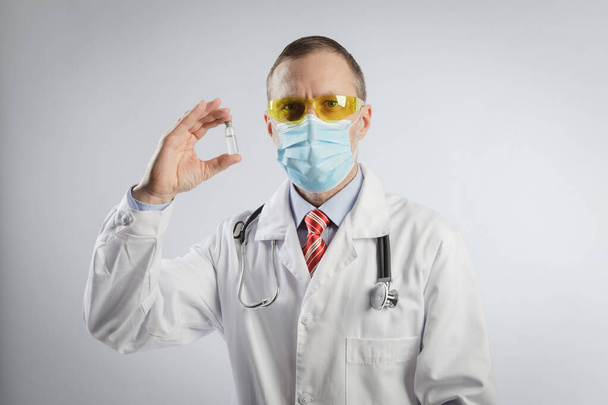 The doctor shows the vaccine in a capsule. Medical specialist in a medical gown, goggles and a mask and a stethoscope on his shoulders - 写真・画像