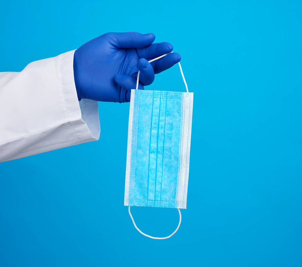 Arzt im weißen Mantel, blaue Latex-sterile Handschuhe, in der Hand medizinische Einmalmasken aus Textil auf blauem Grund, zusätzlicher Schutz vor Viren während einer Epidemie, Pandemie - Foto, Bild