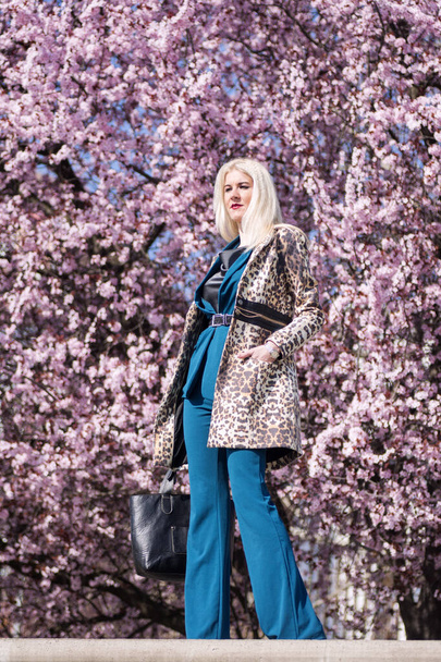 Le modèle féminin aux cheveux blonds séduisant dans des vêtements élégants pose à l'extérieur sur fond d'arbre rose en fleurs
. - Photo, image