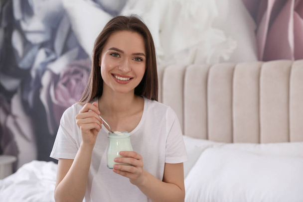Young attractive woman with tasty yogurt on bed at home - Photo, Image