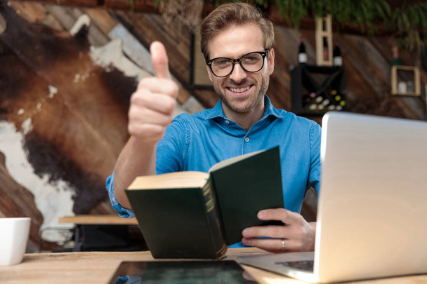 casual άνθρωπος φορώντας γυαλιά κάθεται στο γραφείο και μελετώντας από το βιβλίο, ενώ δίνει ένα αντίχειρες επάνω χαρούμενος στο coffeeshop - Φωτογραφία, εικόνα