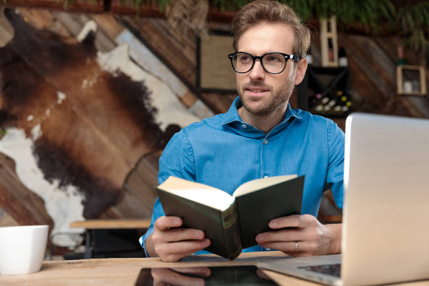 casual man wearing glasses sitting at desk studying from book while looking away happy at the coffeeshop - Photo, Image