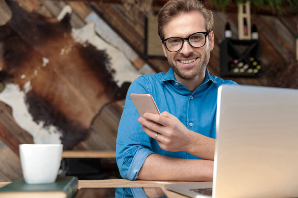 hombre casual con gafas sentado en el escritorio y mensajes de texto de un mensaje en el teléfono mientras mira a la cámara feliz en el coffeeshop
  - Foto, imagen