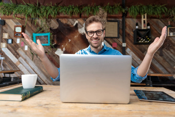 homem casual usando óculos sentado na mesa e trabalhando no laptop enquanto cumprimenta feliz com os braços abertos no coffeeshop
 - Foto, Imagem