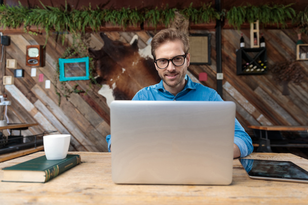 casual man wearing glasses sitting at desk and working on laptop while looking at camera happy at the coffeeshop - Photo, Image