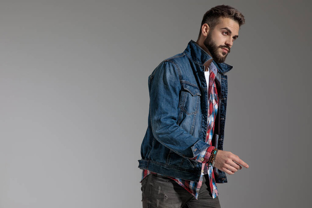 side view of a handsome casual man wearing denim jacket standing and pointing with finger while striking a pose with serious attitude on gray studio background - Zdjęcie, obraz