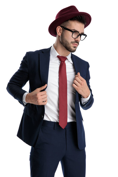 сексуальный бизнесмен в бордовых шляпах стоя и фиксируя куртку с отношением на белом фоне студии
 - Фото, изображение