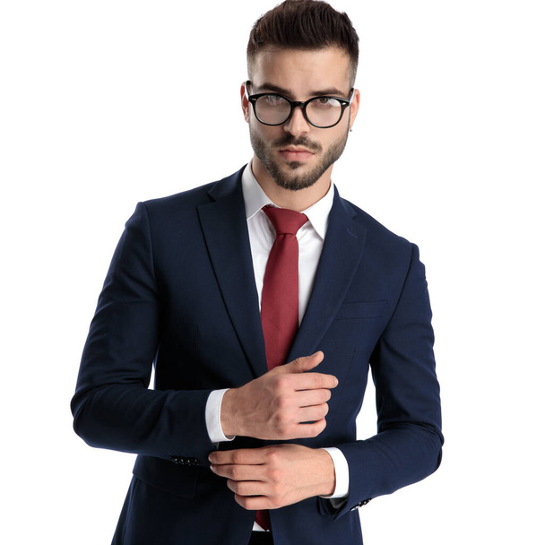 молодий бізнесмен в окулярах стоїть і регулює рукав, одночасно вражаючи позу з серйозним ставленням до білого студійного фону
 - Фото, зображення