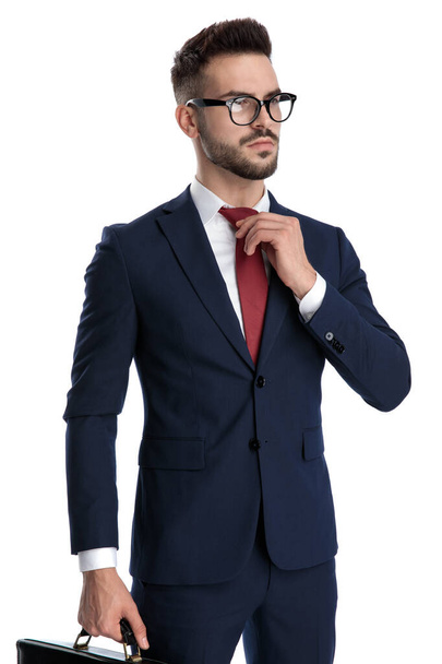 attraktiver Geschäftsmann mit Brille, der eine Aktentasche hält, während er seine Krawatte mit harter Haltung auf weißem Studiohintergrund fixiert - Foto, Bild