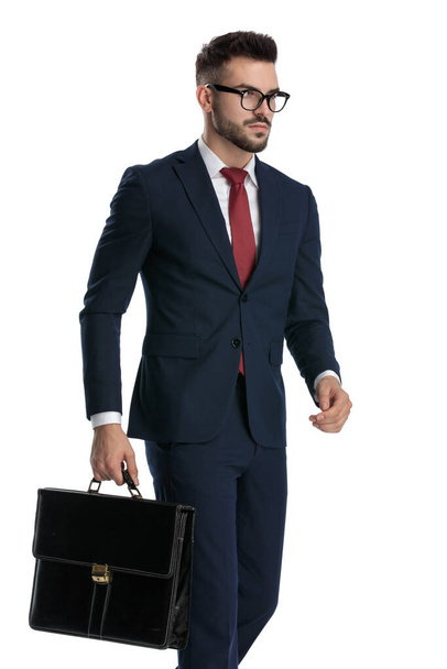 widok z boku przystojny formalny mężczyzna w okularach chodzenie z walizką na rękę i patrząc przed siebie poważnie na białym tle studio - Zdjęcie, obraz