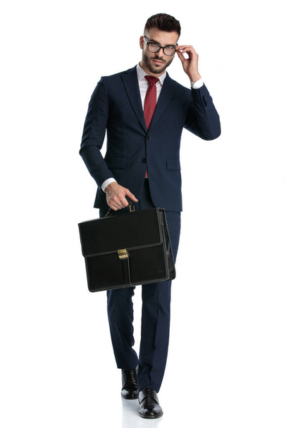 sexy hombre de negocios vistiendo traje azul marino caminando con maletín en la mano y la fijación de sus gafas con estilo en fondo blanco estudio
 - Foto, imagen