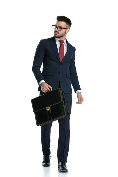 atrakcyjnego biznesmena w okularach, chodzącego z walizką w ręku i odwracającego wzrok na białym tle studia - Zdjęcie, obraz