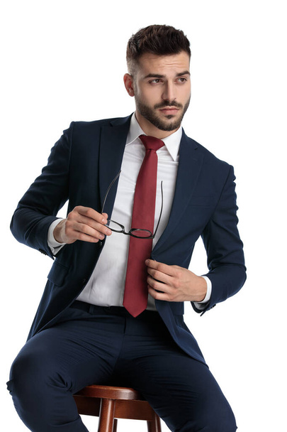 sexy hombre de negocios vistiendo traje azul marino sentado mientras se quita las gafas con actitud fresca y la fijación de la chaqueta en fondo blanco estudio
 - Foto, imagen