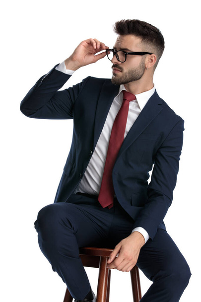 homem de negócios atraente vestindo terno da marinha sentado e consertando seus óculos enquanto olha para o lado sério no fundo do estúdio branco
 - Foto, Imagem