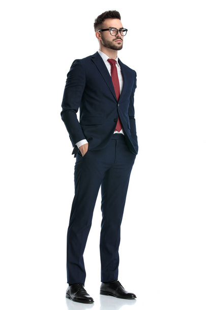 πλαϊνή άποψη ενός όμορφου επιχειρηματία φορώντας γυαλιά στέκεται με τα χέρια στην τσέπη και κοιτάζοντας μπροστά σοβαρό σε λευκό φόντο στούντιο - Φωτογραφία, εικόνα