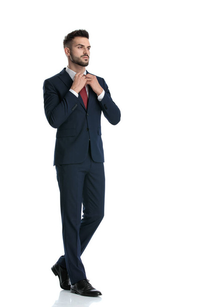 vista laterale di un uomo d'affari attraente che indossa tuta marina a piedi mentre fissa la cravatta con atteggiamento fresco su sfondo bianco studio
 - Foto, immagini