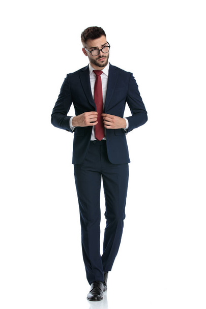 handsome formal man wearing glasses walking while closing his jacket and posing with cool attitude on white studio background - Photo, Image