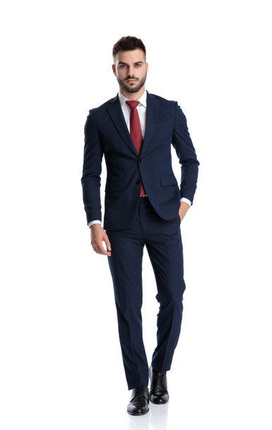 attractive businessman wearing navy suit walking with one hand in pocket and striking a pose with serious attitude on white studio background - Foto, afbeelding
