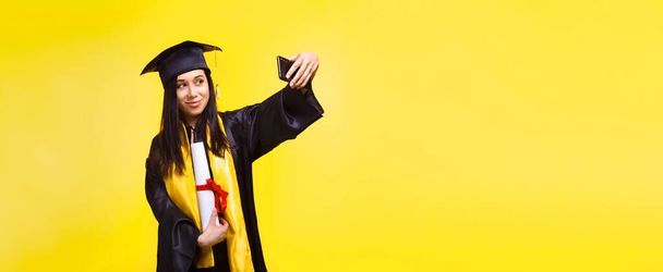 afgestudeerde vrouw maakt selfie foto op telefoon via gele achtergrond, concept van succesvolle voltooiing van studies, panoramische mock-up met plaats voor tekst - Foto, afbeelding