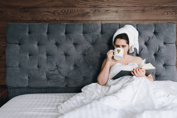 Όμορφη νεαρή καυκάσιος γυναίκα διαβάζει ένα βιβλίο στο κρεβάτι και πίνει ένα φλιτζάνι καφέ κατά τη διάρκεια της καραντίνας λόγω του coronavirus - Φωτογραφία, εικόνα