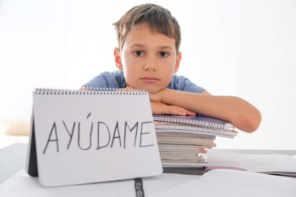 Zmęczony sfrustrowany chłopiec siedzi przy stole z wieloma książkami, ćwiczy książki. Hiszpańskie słowo Auydame - Pomóż mi - jest napisane na otwartym notesie. Trudności w nauce, koncepcja edukacji szkolnej, kwarantanny - Zdjęcie, obraz