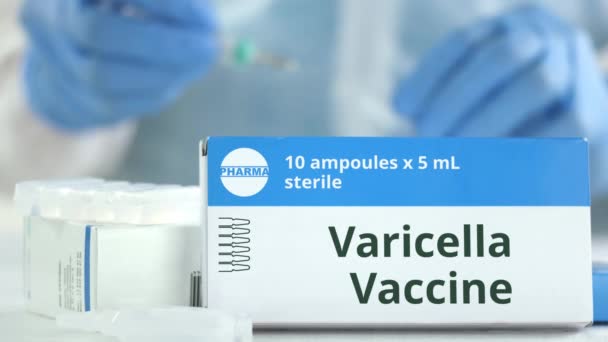 Box with varicella vaccine on the table against blurred lab assistant or doctor. Fictional phaceutical logo - Кадры, видео