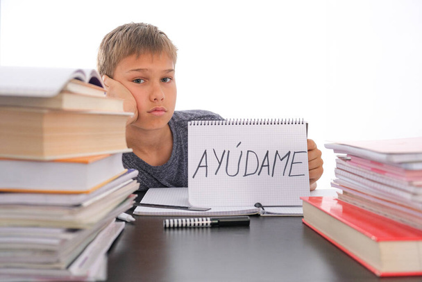 Zmęczony sfrustrowany chłopiec siedzi przy stole z wieloma książkami, ćwiczy książki. Hiszpańskie słowo Auydame - Pomóż mi - jest napisane na otwartym notesie. Trudności w nauce, koncepcja edukacji szkolnej, kwarantanny - Zdjęcie, obraz