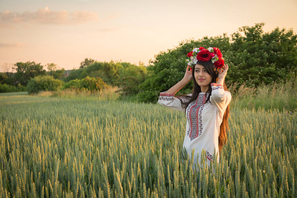 Csinos fiatal nő visel hagyományos ukrán ruhát és virágkoszorú séta a búzamezőn, gyönyörű etnikai lány kézzel készített díszített virágos korona csodálja a természetet. Arany óra. - Fotó, kép