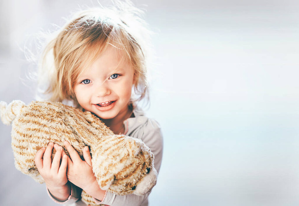 Крупный план милой блондинки с голубыми глазами, держащей плюшевую игрушку в руке и улыбающейся невинно копирующей пространство
 - Фото, изображение