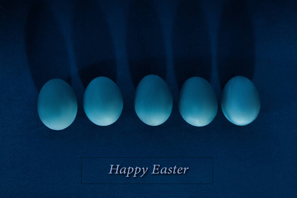 5 ovos azuis de pé sobre um fundo uniforme e lançar uma sombra dura de volta isolado, sobre um fundo azul escuro, flat lay. Conceito de férias de Páscoa e primavera. Espaço de cópia, vista superior
. - Foto, Imagem