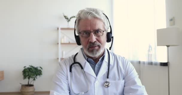 Senior doctor wears headset makes distant video call, webcam view - Imágenes, Vídeo