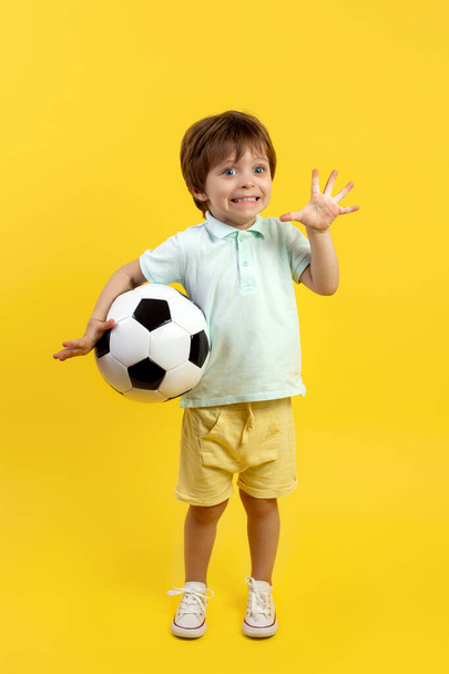 Adorable little kid in casual summer outfit holding a soccer ball and posing with funny face over yellow background. - Fotó, kép