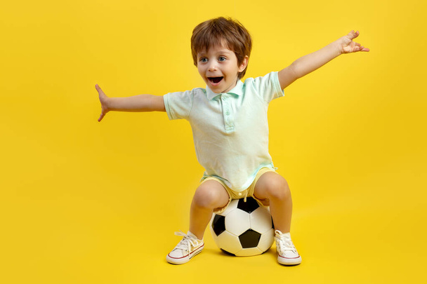 Cute little boy in casual summer outfit sitting on the soccer ball and stretching out arms over yellow background. - 写真・画像