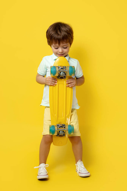 Uroczy chłopczyk w luźnym stroju letnim trzymający pennyboard lub deskorolkę na żółtym tle. - Zdjęcie, obraz