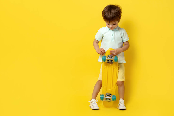 Cute little boy in casual summer outfit holding pennyboard or skateboard over yellow background. - Φωτογραφία, εικόνα