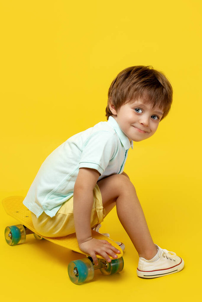 Charming little boy in casual summer outfit sitting on pennyboard or skateboard over yellow background. - 写真・画像