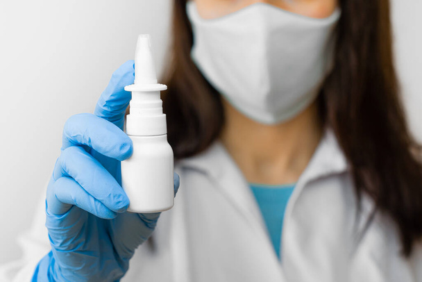 Een vrouw die neusspray gebruikt voor griepbehandeling of loopneus houdt medicijnen in de hand. Dokter in beschermende masker en blauwe handschoenen toont neusspray voor de behandeling van ziekten - Foto, afbeelding