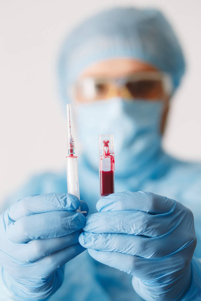 Κοντινό πλάνο της σύριγγας που κρατά το χέρι του γιατρού και δείγμα αίματος. Ιατρικός εξοπλισμός. Εξέταση αίματος. Ένας γιατρός που φοράει μέσα ατομικής προστασίας, συμπεριλαμβανομένων μάσκα, goggle, και κοστούμι για την προστασία COVID 19 coronavirus λοίμωξη. - Φωτογραφία, εικόνα