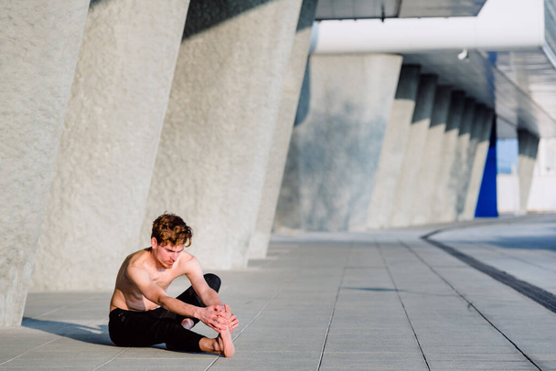 Молодой рыжеволосый спортсмен делает растяжку после тренировки, чтобы получить гибкость и расслабить мышцы, на открытом воздухе с несосредоточенным фоном. - Фото, изображение