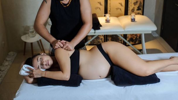 Myöhemmin näkymä raskaana olevasta valkoihoisesta nuoresta naisesta, joka makaa sängyllä ja jolla on rentouttava itämainen raskaudenaikainen hieronta käsivarsillaan.
 - Valokuva, kuva