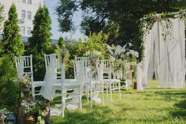 Rows of white empty chairs on a lawn near a wedding round arch decorated with white curtains and flowers, close to trees and a building, before a wedding ceremony  - Photo, Image